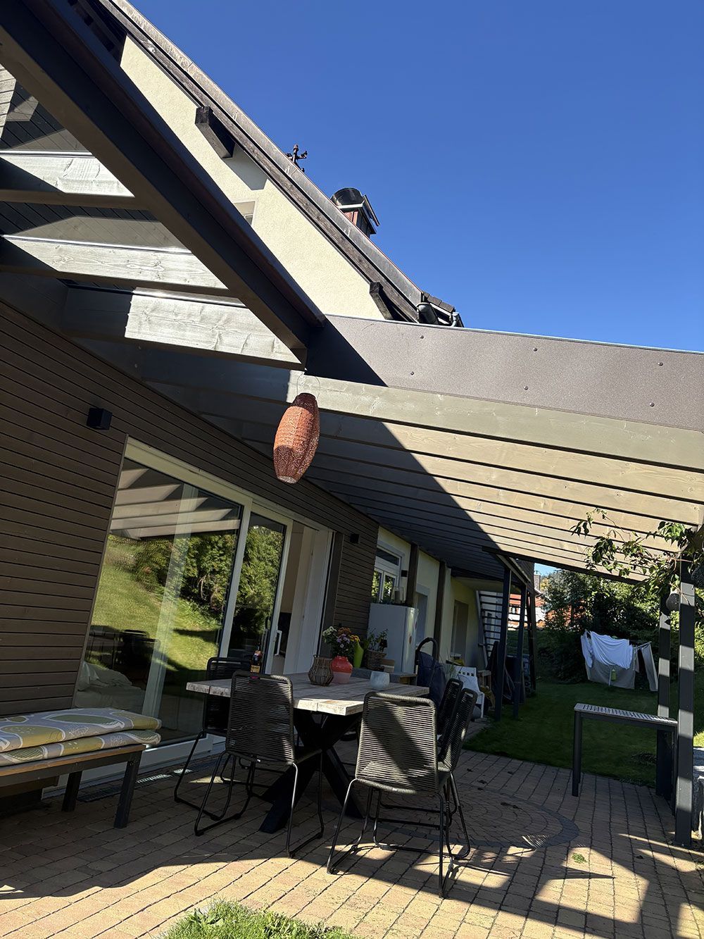 Eine Terrasse unter einer hölzernen Pergola an einem sonnigen Tag, ausgestattet mit einem Tisch, Stühlen und einer hängenden dekorativen Laterne.