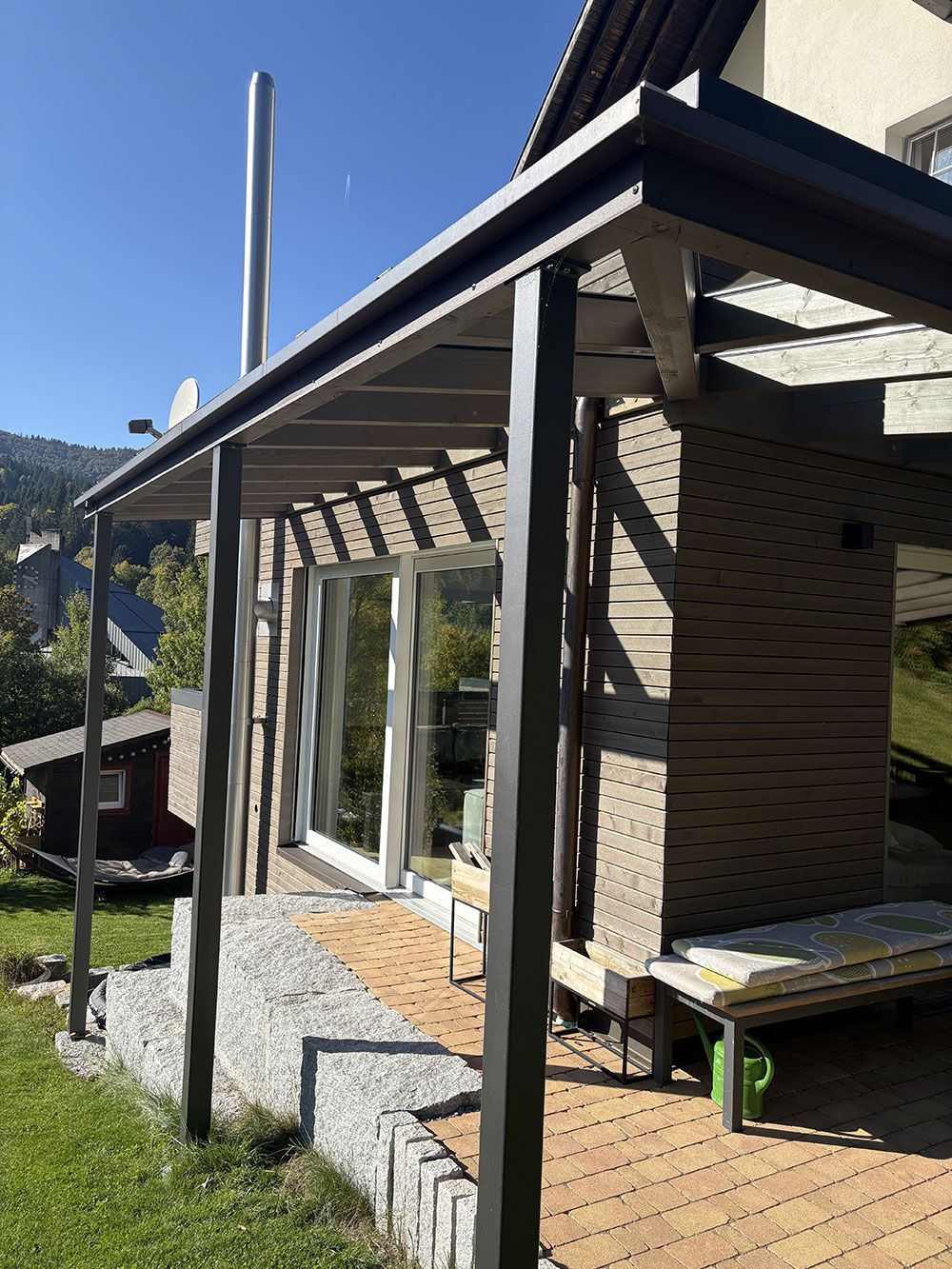 Eine moderne hölzerne Terrassenüberdachung mit schwarzem Rahmen und mit Kies gefüllten Stützpfeilern grenzt an ein Haus mit Glastüren.