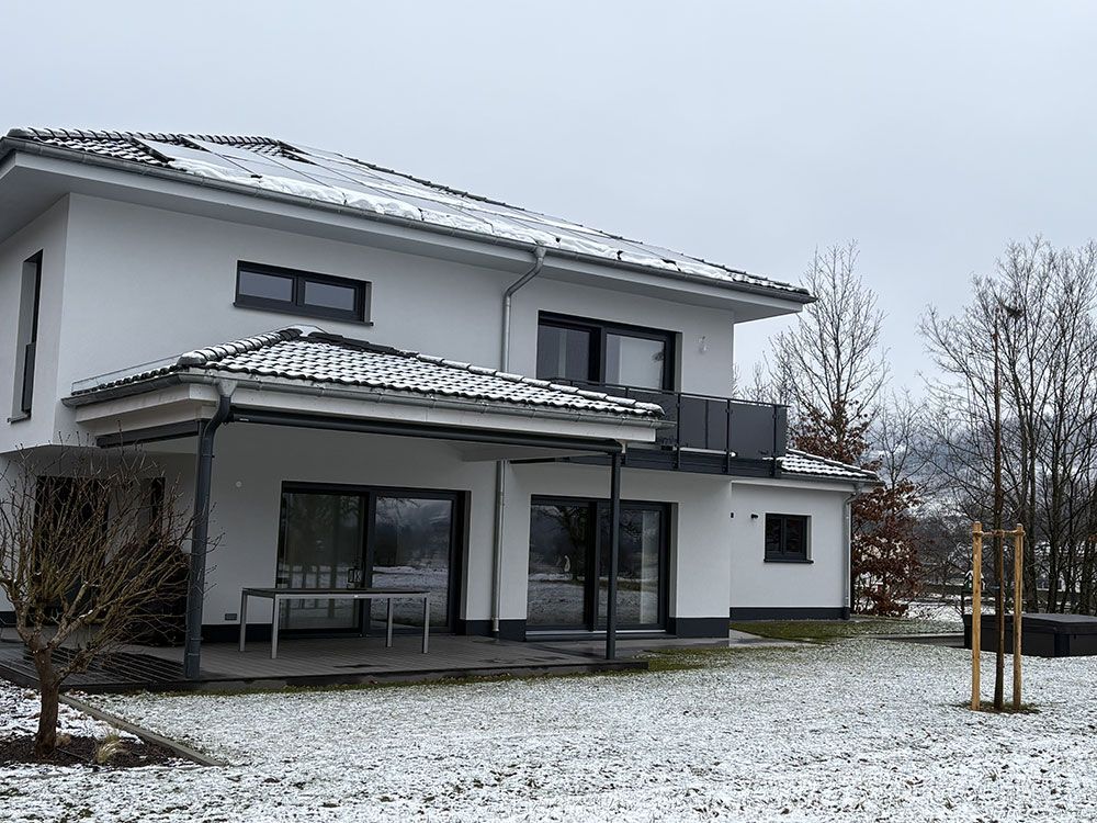 Ein modernes, zweistöckiges weißes Haus mit überdachter Terrasse und dunklen Fensterrahmen, eingebettet in einen leicht schneebedeckten Garten.