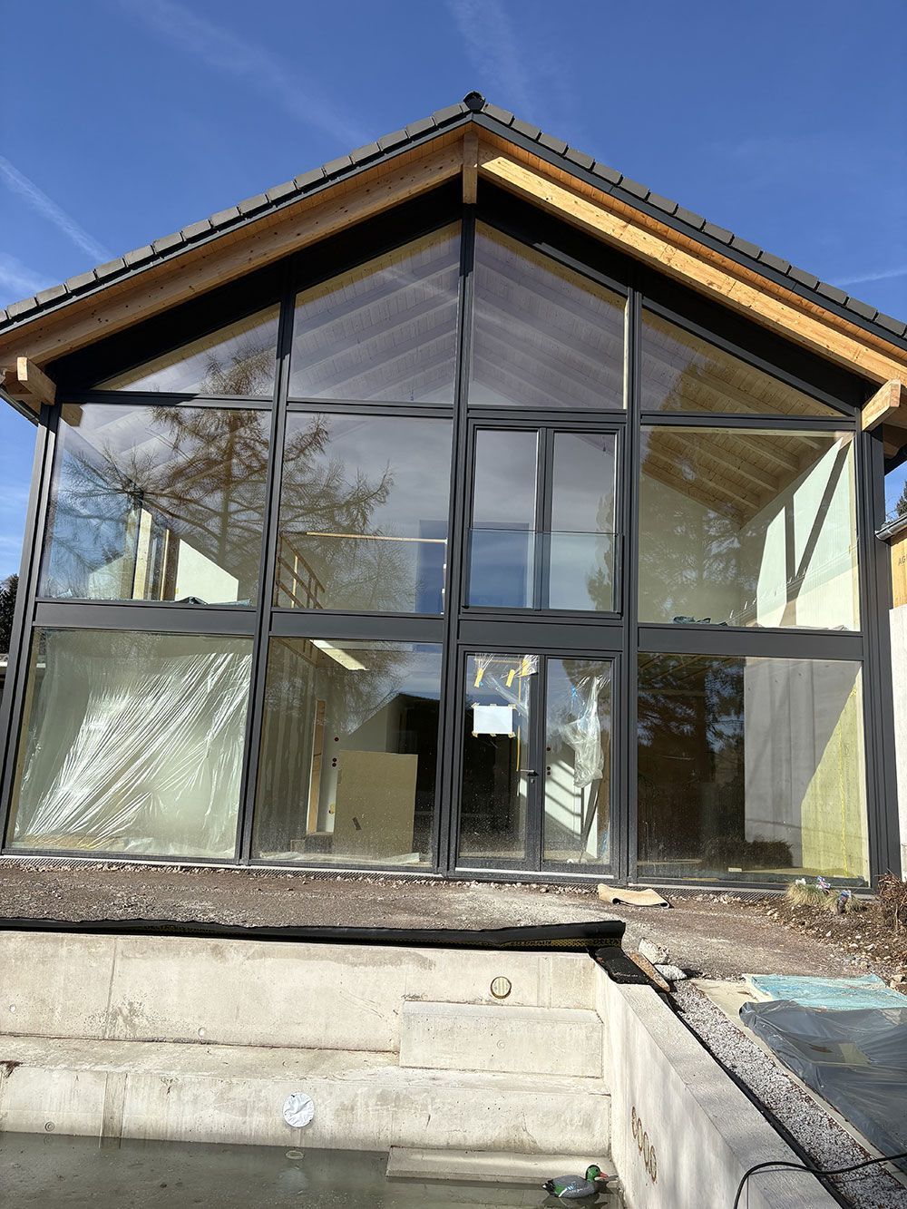 Ein modernes, im Bau befindliches Gebäude mit großer Glasfassade, hölzernen Dachbalken und einem unfertigen Beton-Terrassenbereich.