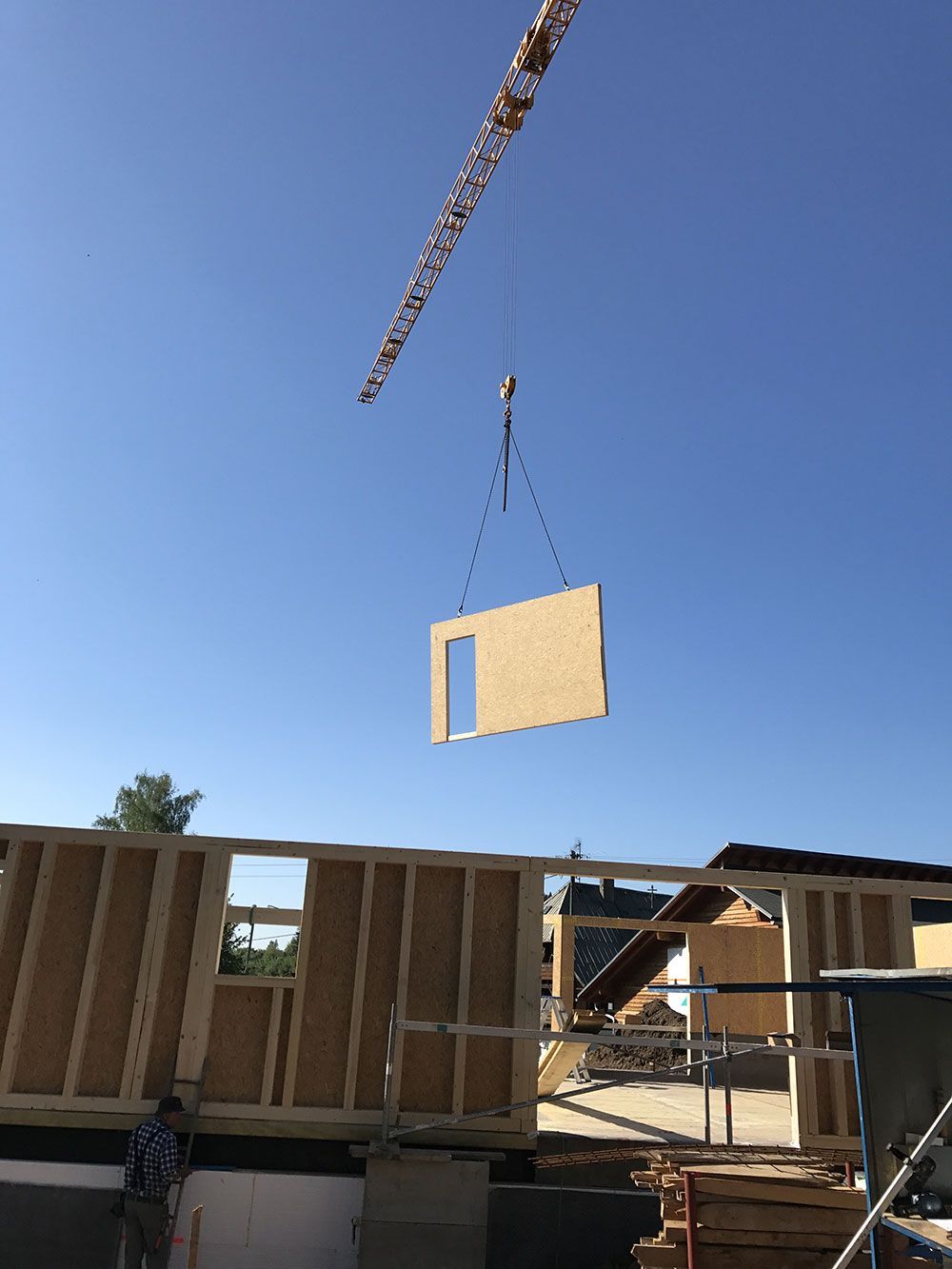 Bei klarem Himmel senkt ein Kran eine große, vorgefertigte Holzwandplatte auf das Gerüst eines im Bau befindlichen Hauses.