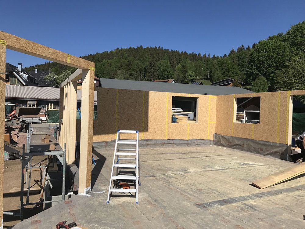 Eine Baustelle, auf der unter strahlend blauem Himmel hölzerne Wandpaneele auf einem Betonfundament errichtet werden.
