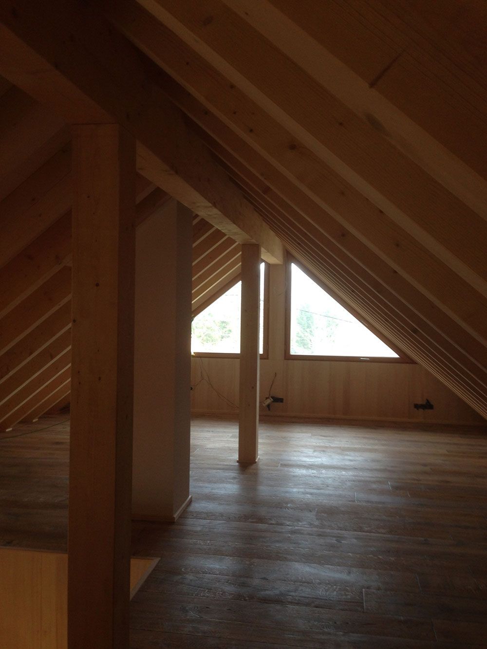 Ein unfertiges Dachzimmer mit freiliegenden Holzbalken, hellem Holzboden und zwei dreieckigen Fenstern.