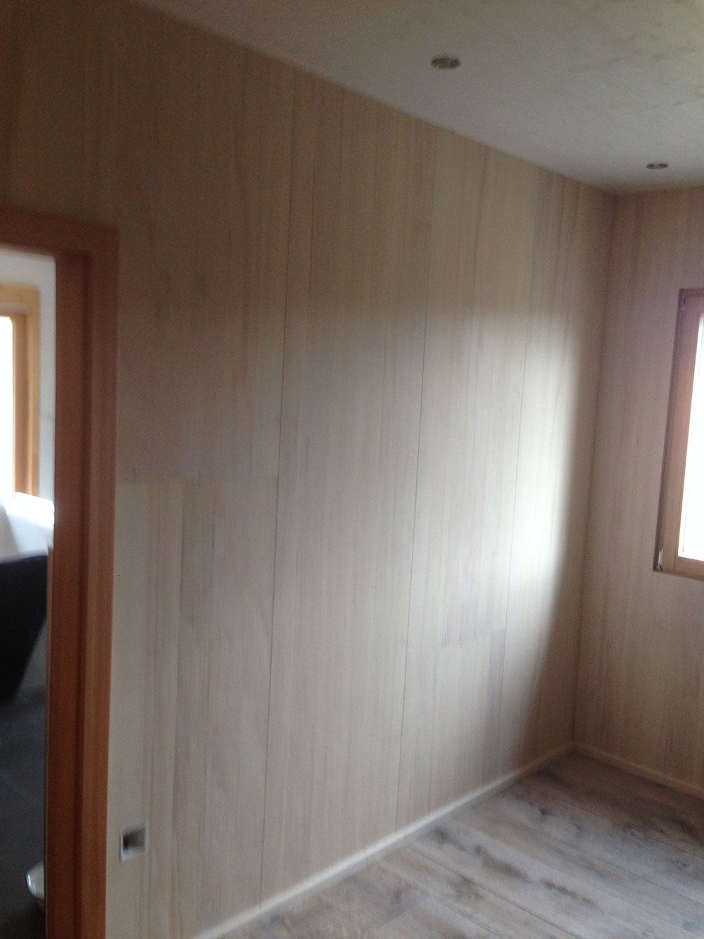 Ein Zimmer mit hellen, holzgetäfelten Wänden, einem Türrahmen aus Holz, einem Fenster und einem Boden in Holzoptik.