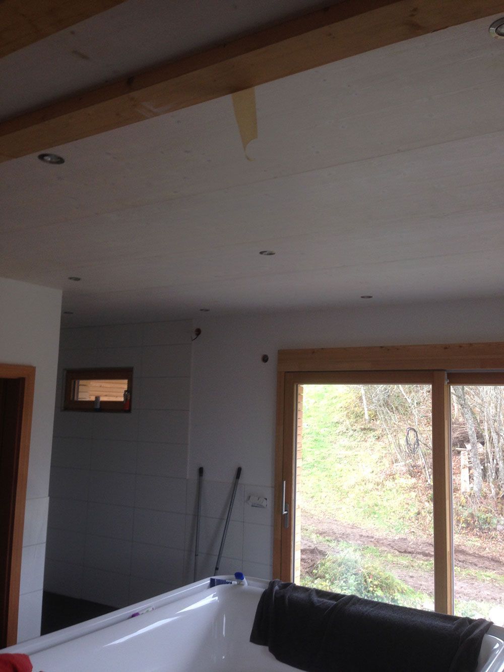 Ein Innenraum mit einer Badewanne im Vordergrund, weißen Wänden, Deckeneinbaustrahlern, Holzbalken und einem großen Fenster.