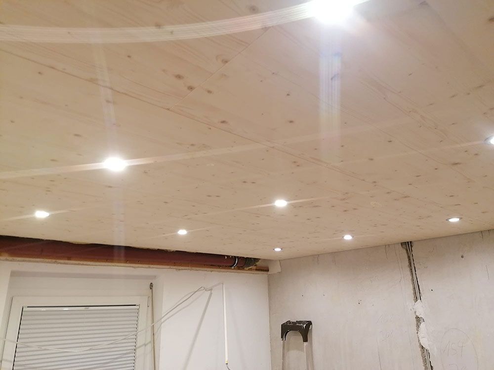 Über einem Raum mit unfertigen Wänden und einem Fenster befindet sich eine im Bau befindliche Holzdeckenplatte mit eingebauten Einbaustrahlern.