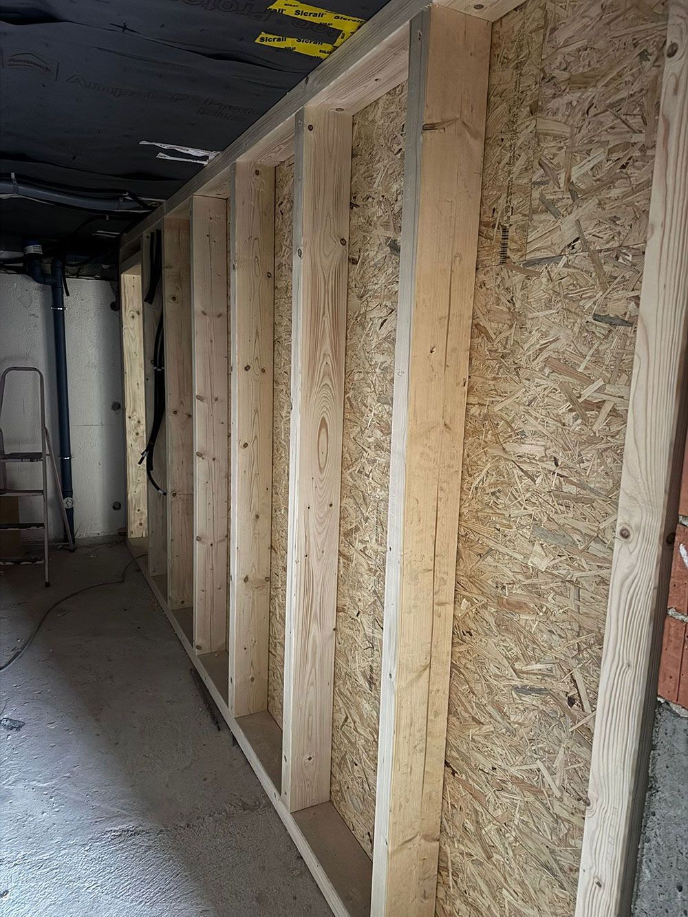 Eine im Bau befindliche Holzrahmenwand mit OSB-Platten in einem unfertigen Keller mit freiliegender schwarzer Deckenisolierung.