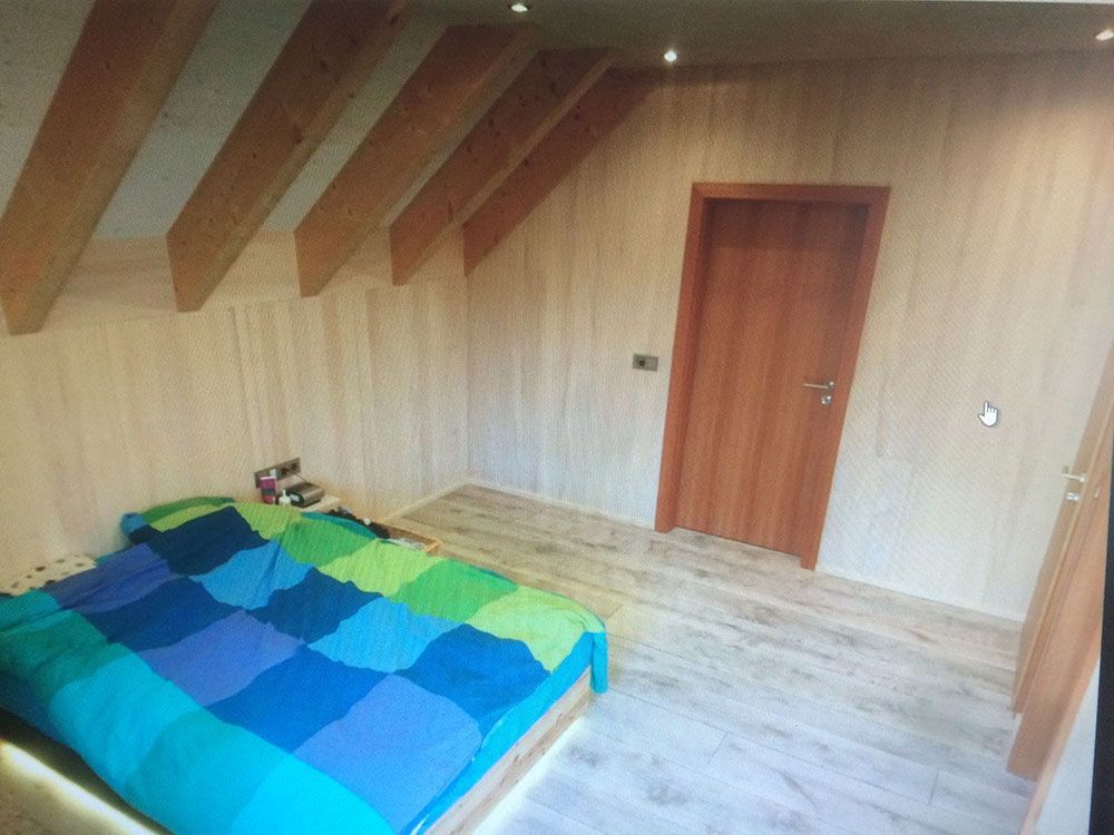 Ein Schlafzimmer mit schräger Holzdecke, einem Bett mit blau-grüner Steppdecke und einer hellen Holztür an einer hellen Wand.