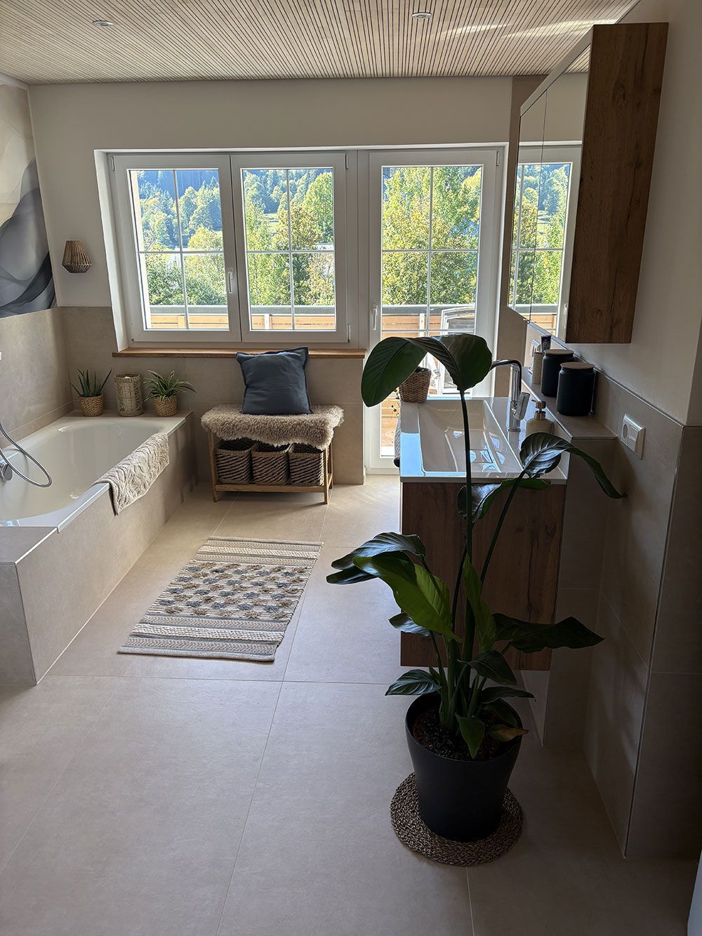 Ein helles Badezimmer mit Badewanne, einem Waschtisch aus Holz, einer hohen Topfpflanze und einem Fenster mit Blick auf eine grüne Landschaft.