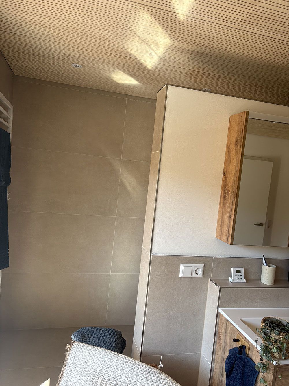 Ein modernes Badezimmer mit strukturierter Decke, großen beigefarbenen Wandfliesen, einem Waschtisch aus hellem Holz, einem Spiegel und einer kleinen Topfpflanze.