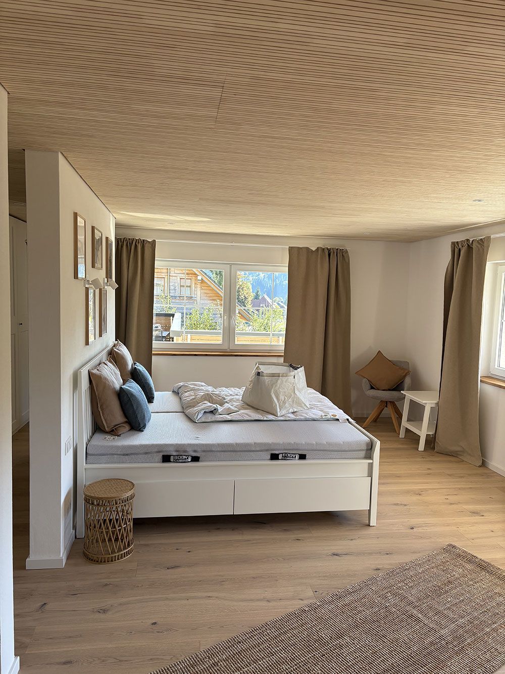 Ein helles Schlafzimmer mit weißem Bett, beigefarbenen Vorhängen, strukturierter Decke und Holzboden, gesehen von einer Türöffnung aus.