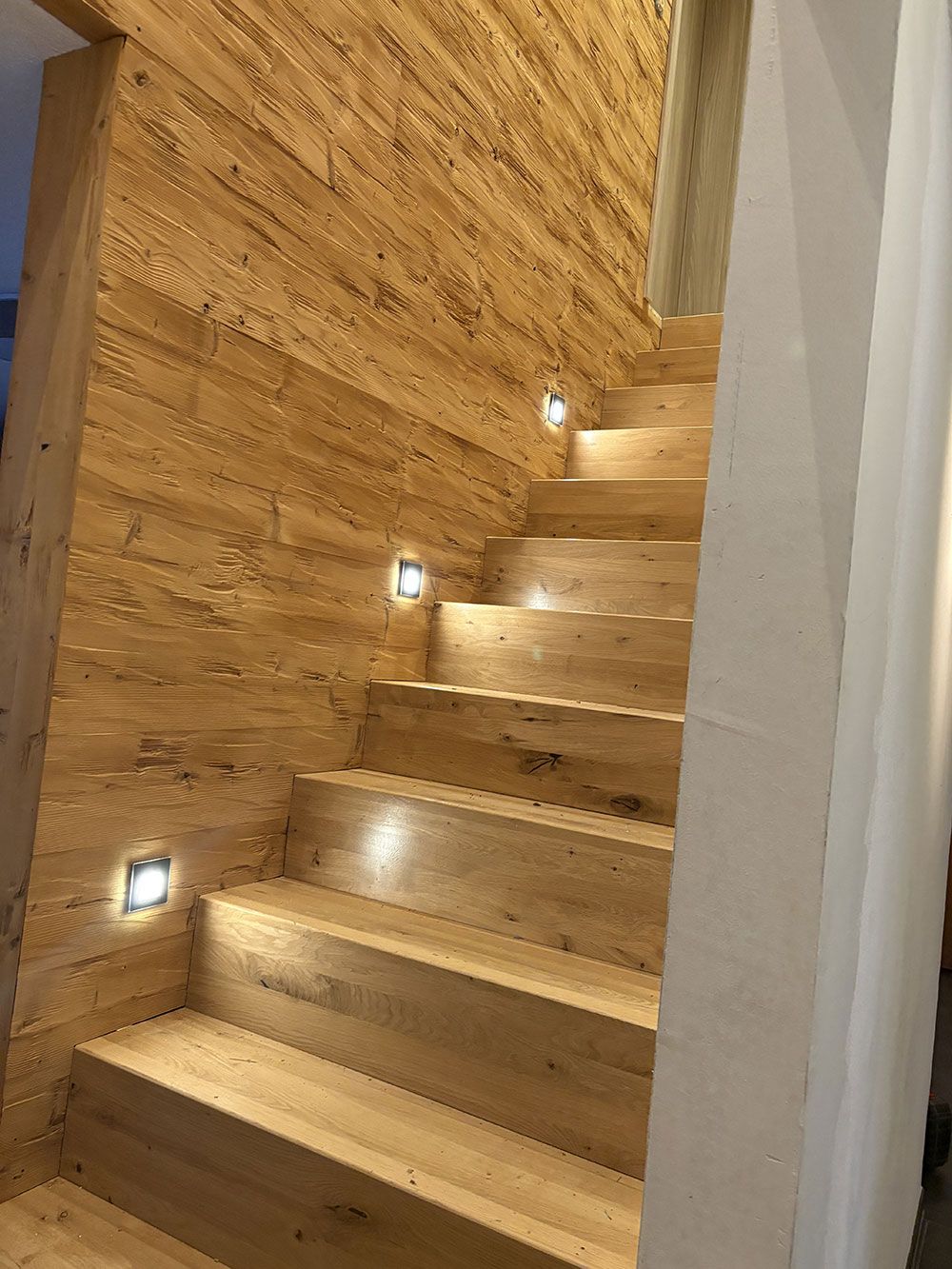 Eine Holztreppe, die von kleinen quadratischen LED-Wandleuchten beleuchtet wird, die in die strukturierte, holzvertäfelte Wand eingelassen sind.