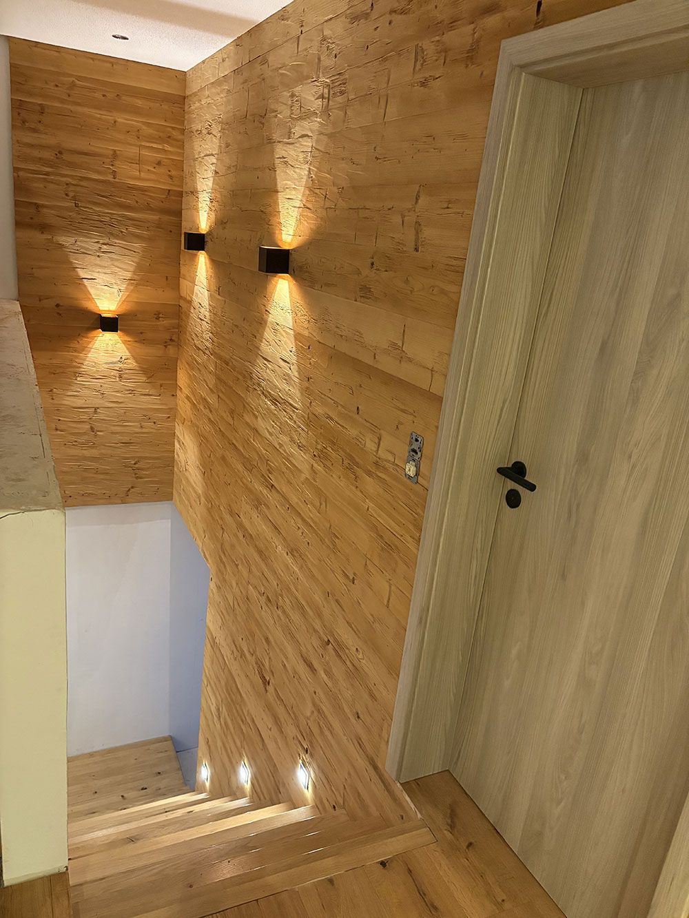 Treppenhaus mit hellen Holzwänden und -böden, beleuchtet durch moderne schwarze Wandleuchten und eingelassene Stufenleuchten.