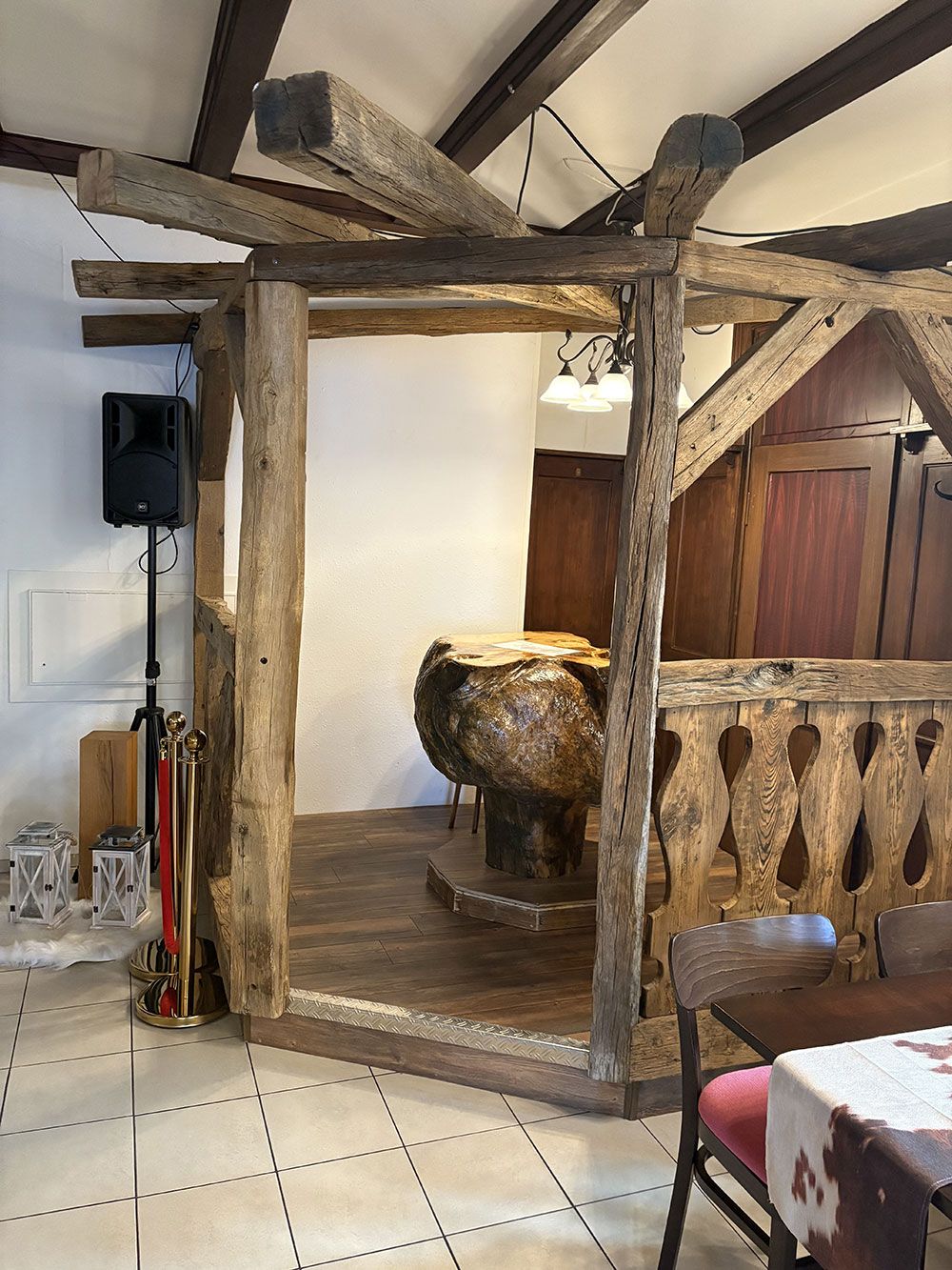 Ein rustikaler Holzpavillon in einem Raum mit Fliesenboden, mit einem geschnitzten Holzsockel und einem dekorativen Geländer.