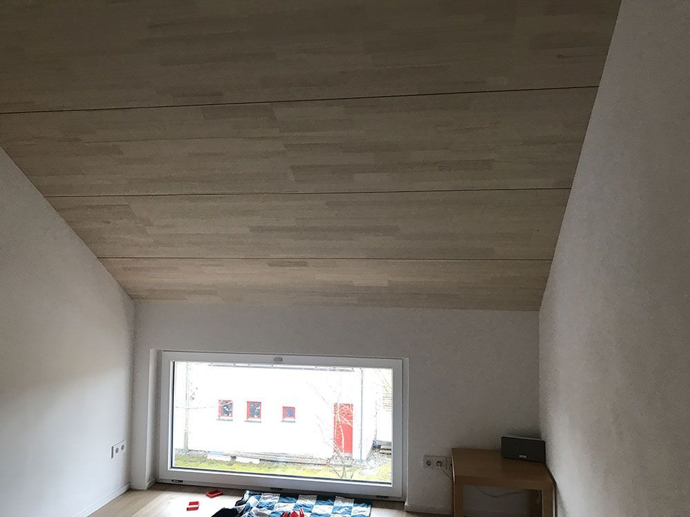Ein Dachzimmer mit heller, holzgetäfelter, schräger Decke, weißen Wänden und einem großen Fenster mit Blick auf ein Gebäude.