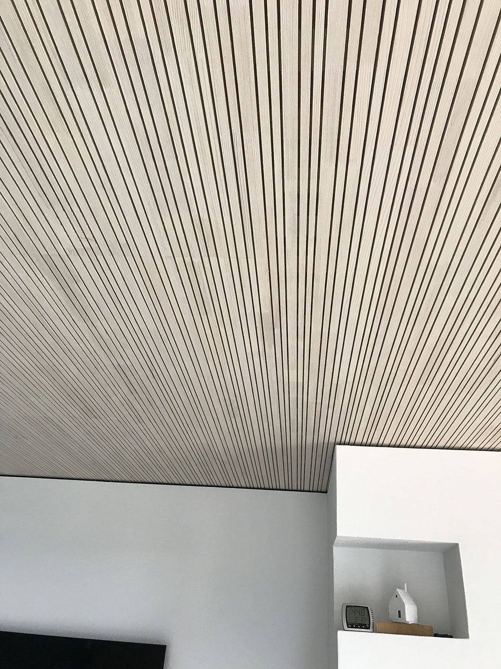 Eine mit strukturierten, hellen Holzlamellen verkleidete Decke über einer schlichten weißen Wand mit einem kleinen eingelassenen Regal.