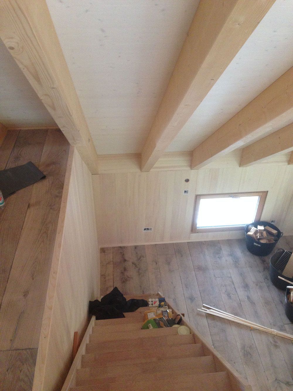 Blick von einer hölzernen Treppe im Dachgeschoss hinunter in einen Raum mit hellen Wänden, Holzfußboden und einem kleinen Fenster.