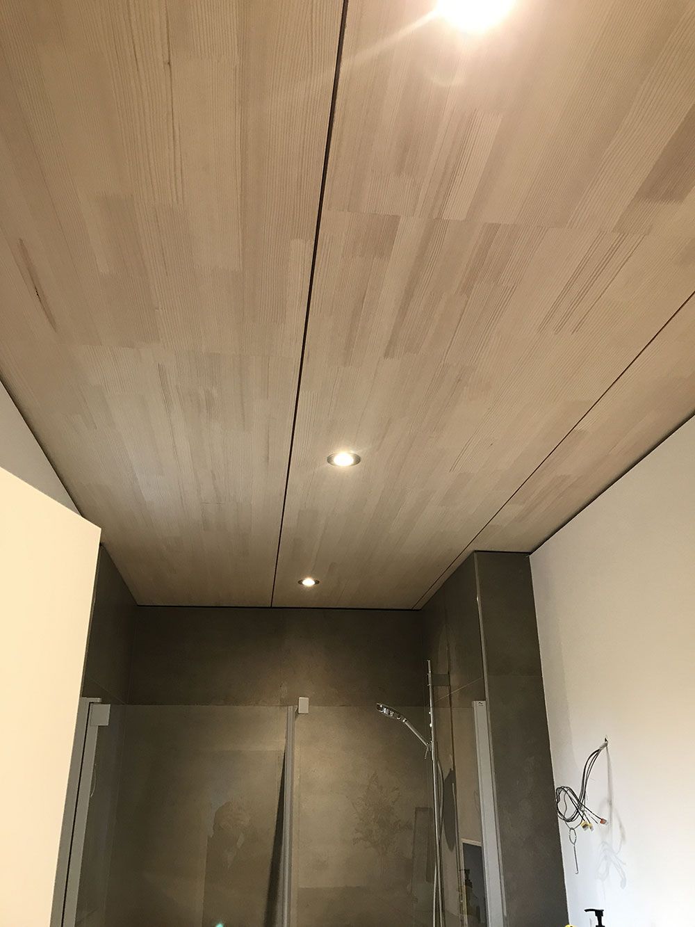 Eine Ansicht aus der Froschperspektive auf eine Badezimmerdecke mit hellen Paneelen in Holzoptik, Einbauleuchten und darunter liegenden dunklen Fliesenwänden.