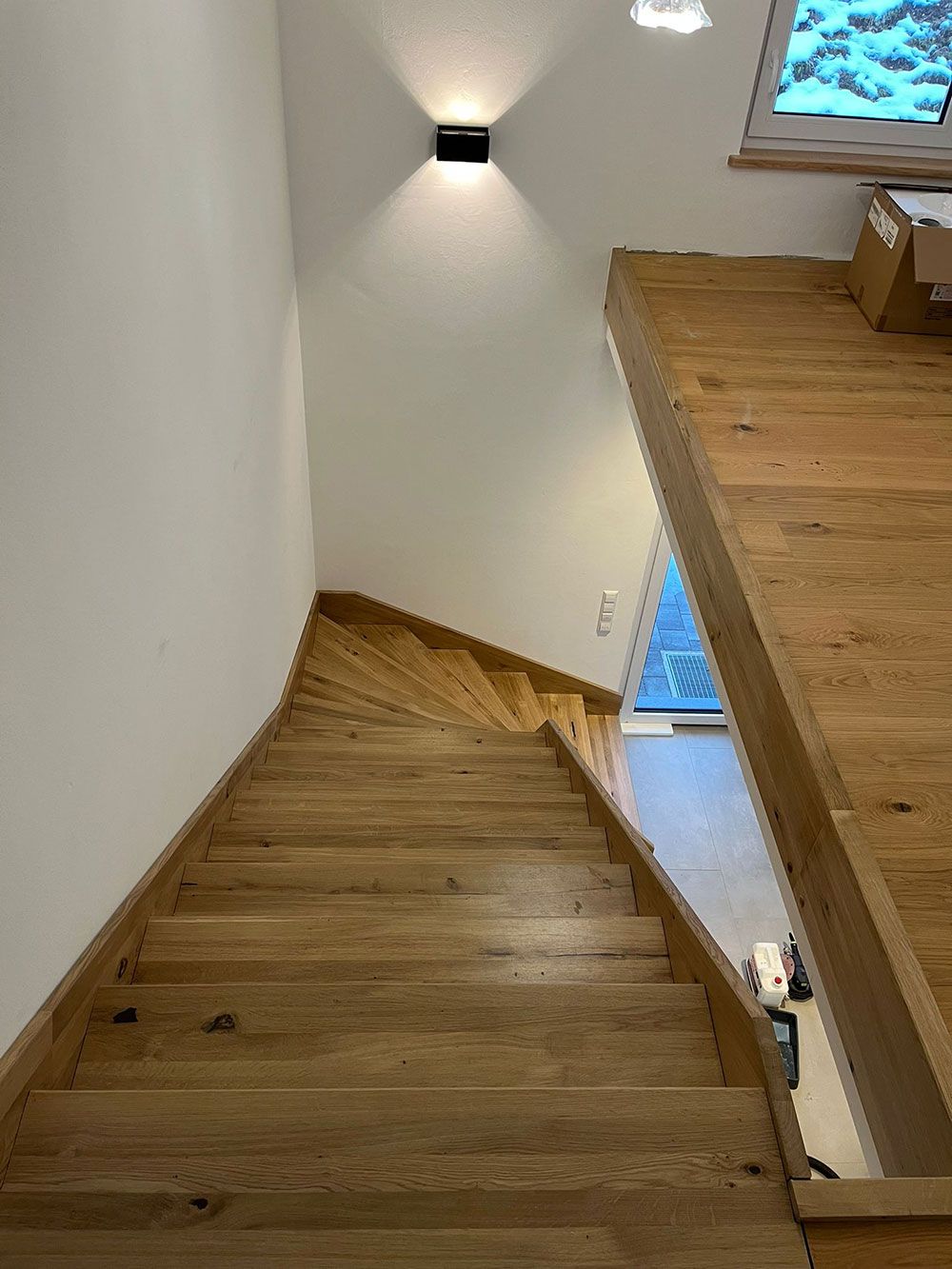 Blick von oben auf eine Holztreppe in einem modernen Haus mit weißen Wänden und einer Wandleuchte.