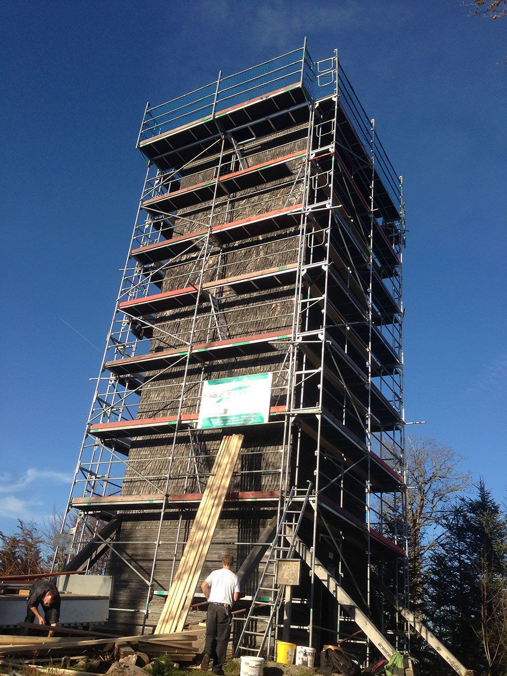Ein hoher Steinturm, umgeben von einem mehrstöckigen Gerüst, vor einem klaren blauen Himmel; am Fuß des Turms sind zwei Arbeiter zu sehen.