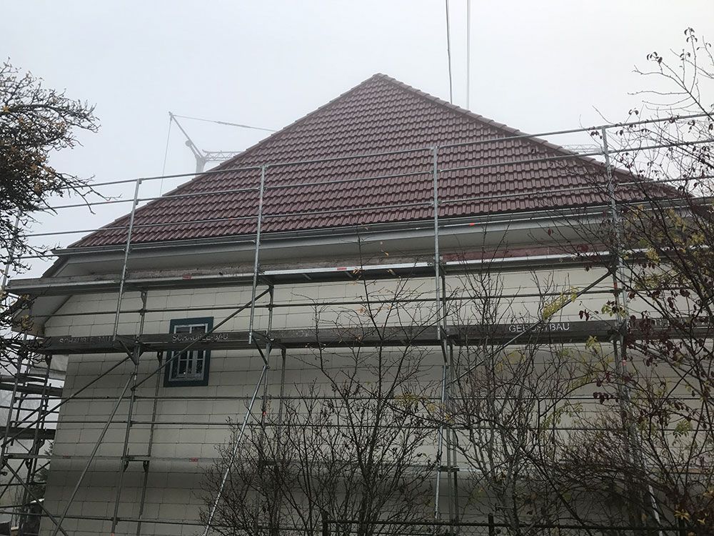 Ein Gebäude mit einem Walmdach, das mit roten Ziegeln gedeckt ist und von einem Metallgerüst für Außensanierungsarbeiten umgeben ist.