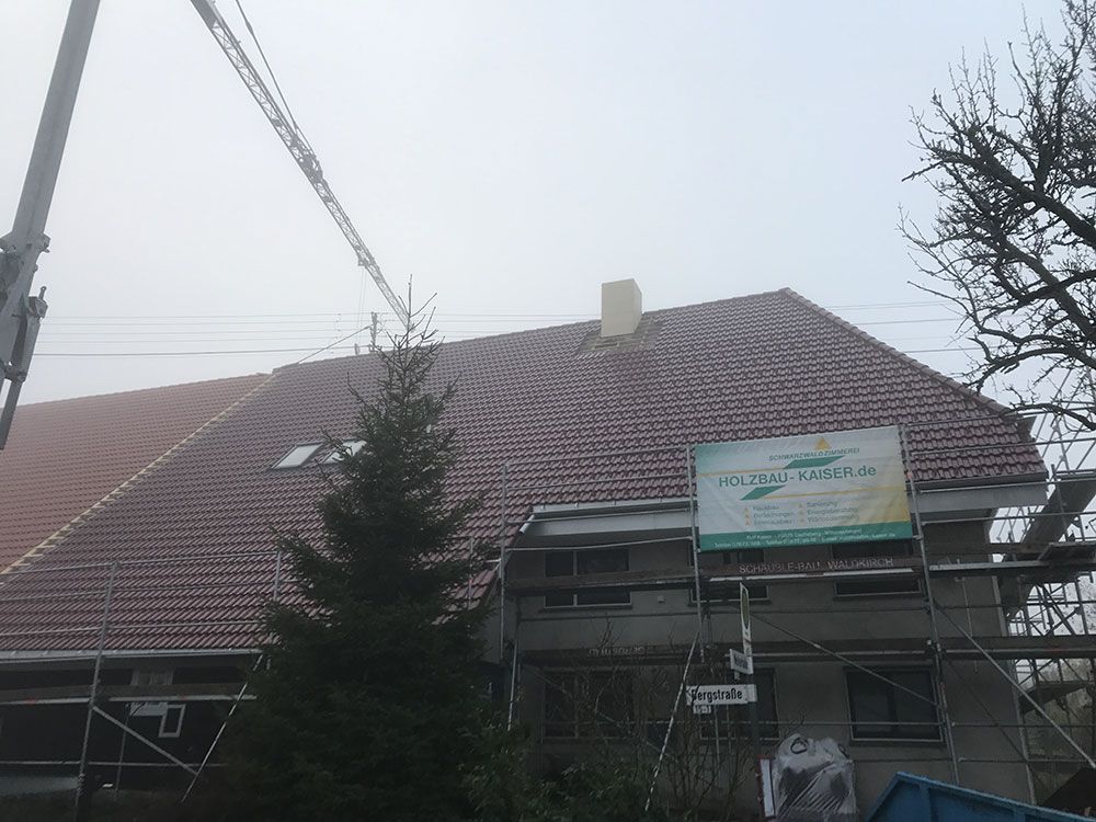 Ein Haus, das gerade renoviert wird, mit Gerüsten und einem grünen Schild, mit rotem Ziegeldach und Schornstein unter grauem Himmel.