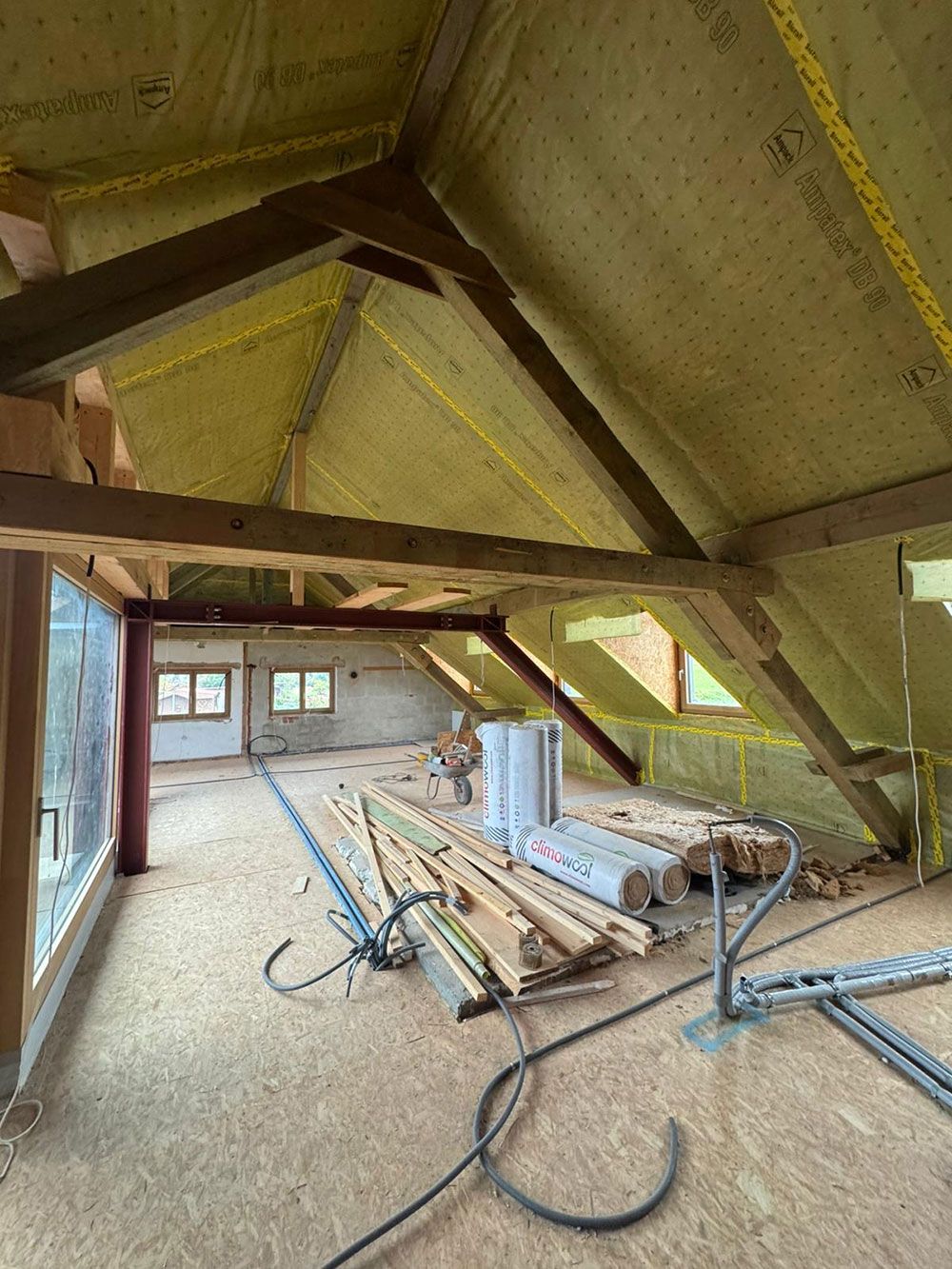 Dachbodenausbau mit freiliegenden Holzbalken, gelben Wärmedämmplatten an den Dachsparren und verstreuten Baumaterialien.