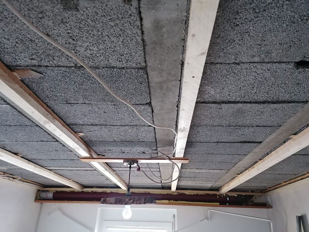 Eine Ansicht aus der Froschperspektive auf eine im Bau befindliche Decke mit freiliegenden grauen Betonblöcken, Holzbalken und einer hängenden Glühbirne.