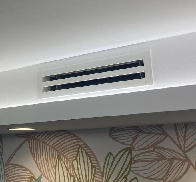 Grille de ventilation rectangulaire blanche dans un plafond blanc, avec un mur décoratif à motifs floraux au premier plan.