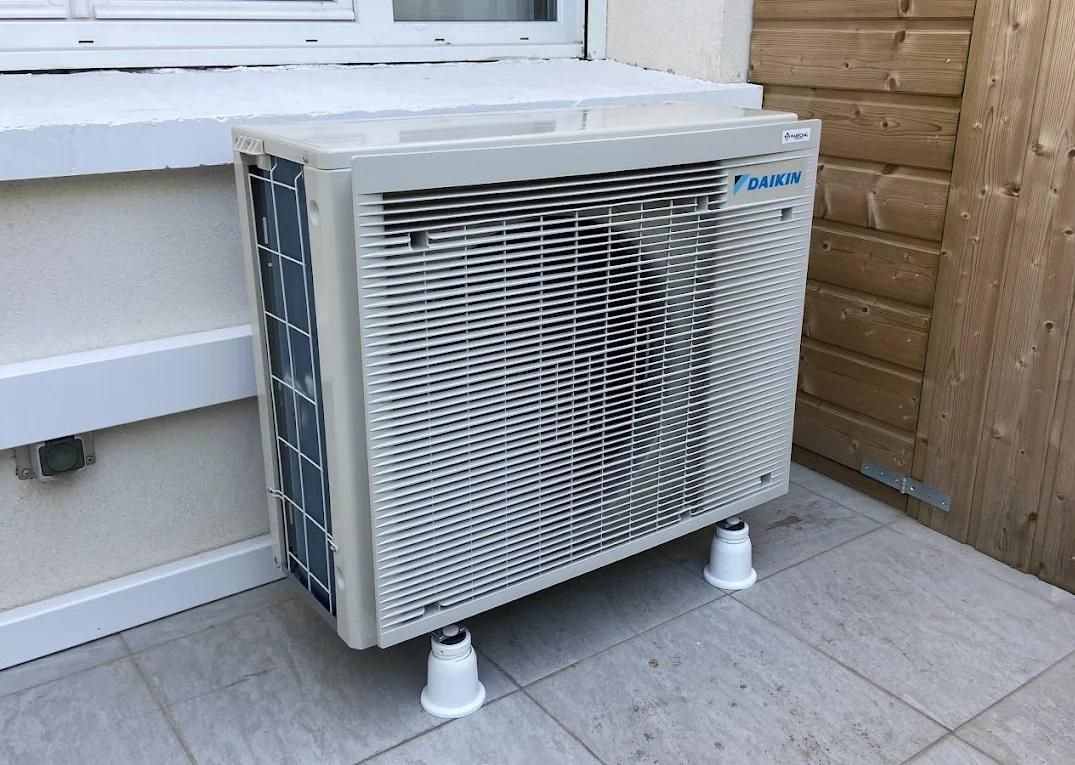 Un climatiseur extérieur sur des supports blancs, placé à côté d'un mur, d'une fenêtre et d'une clôture en bois.