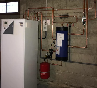 Une pompe à chaleur Daikin blanche et des tuyaux en cuivre sont installés sur un mur de sous-sol en béton, avec un cylindre bleu et un vase d'expansion rouge.