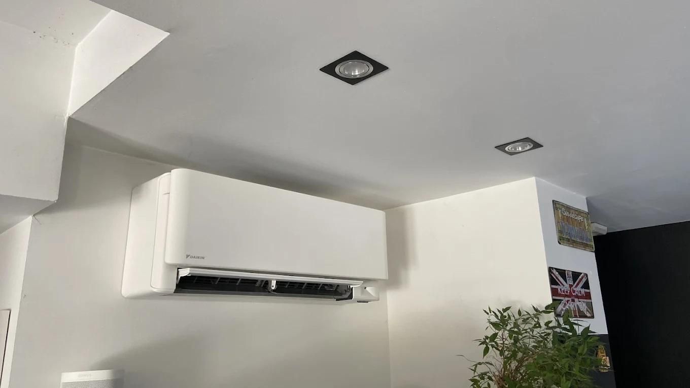 Unité de climatisation blanche sur un mur blanc, avec des spots encastrés au plafond et une plante en pot à proximité.