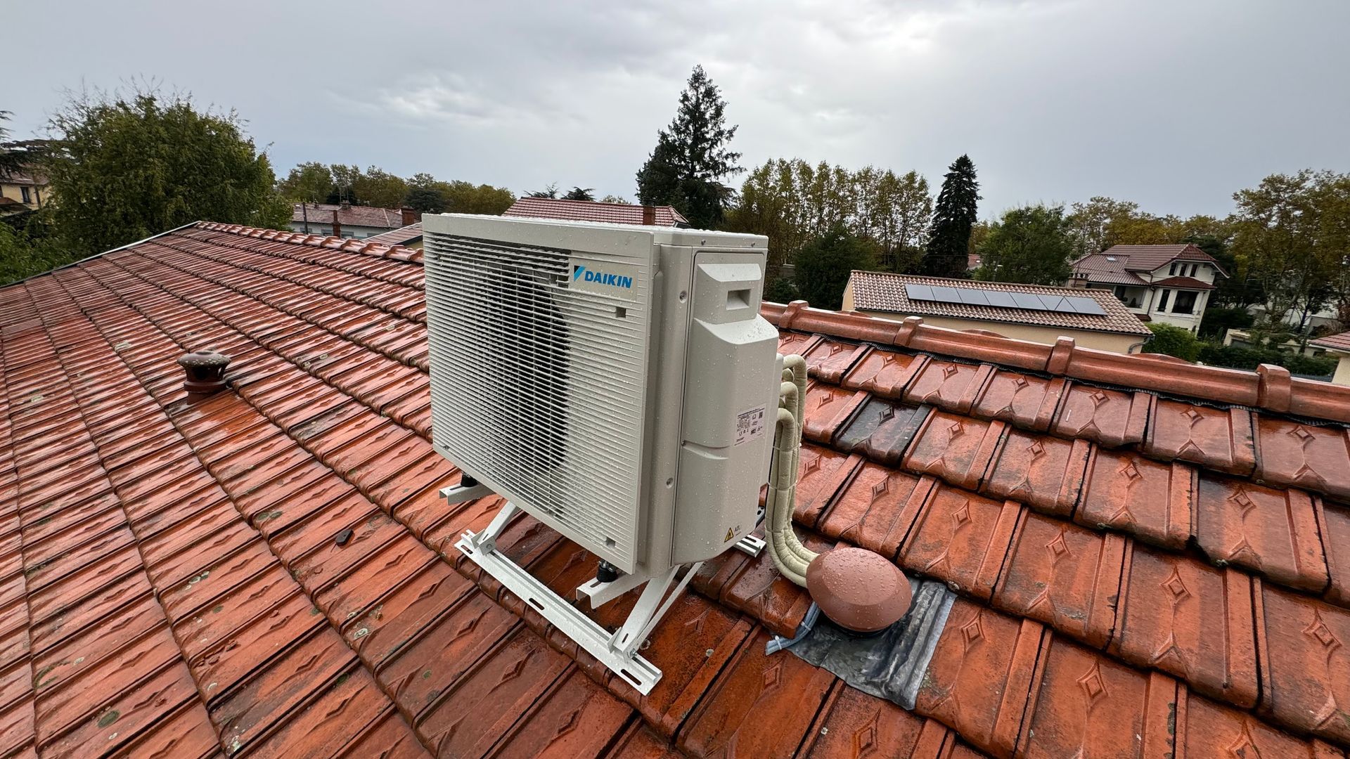 Unité de climatisation sur un toit de tuiles rouges ; ciel nuageux.