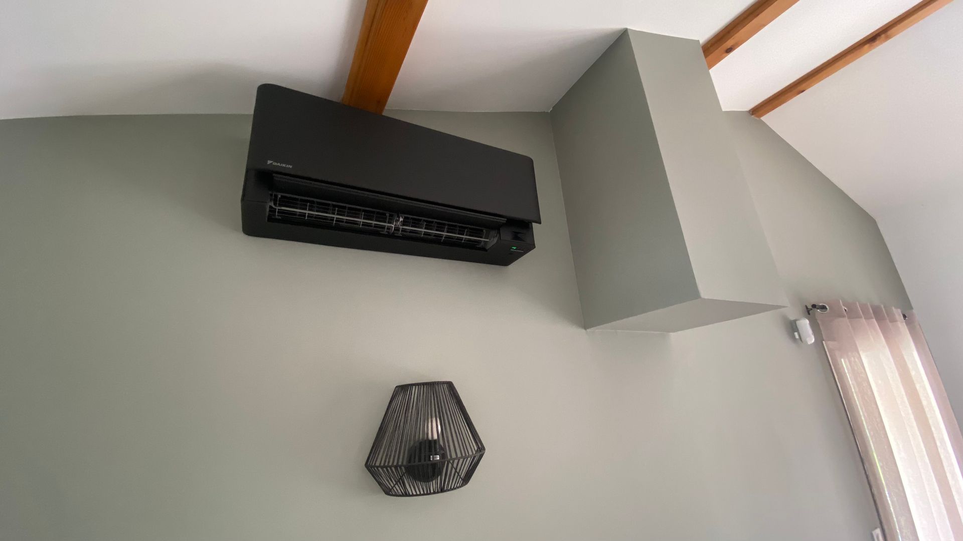 Unité de climatisation murale noire sur un mur vert pâle, sous des poutres en bois.