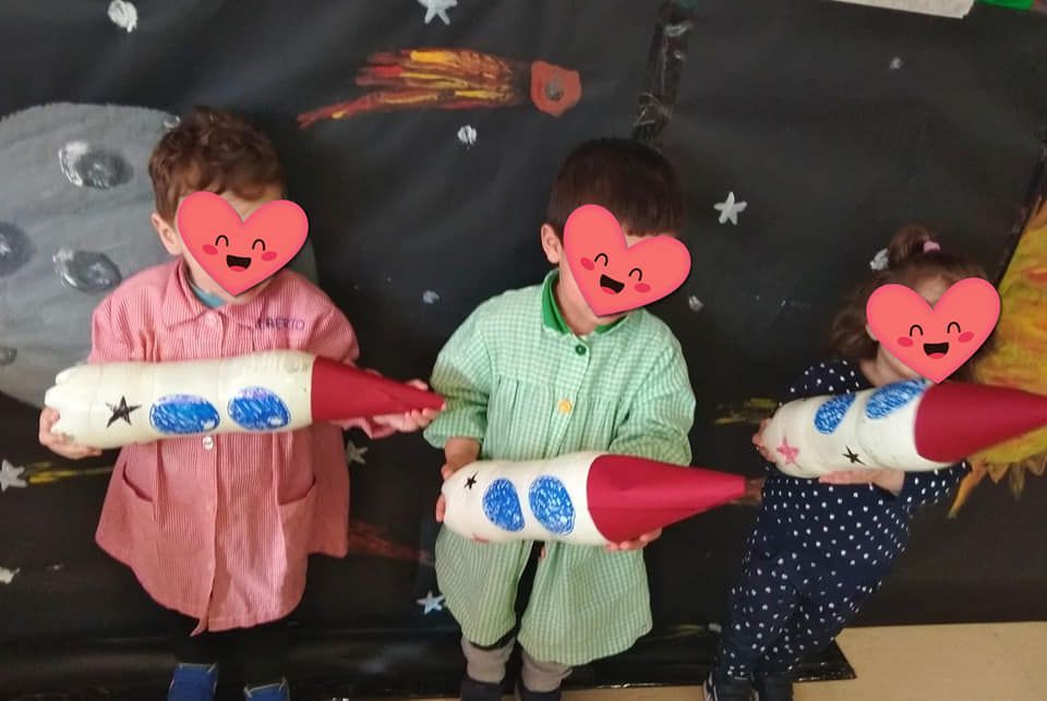 Tres niños sostienen cohetes de papel frente a una pared.