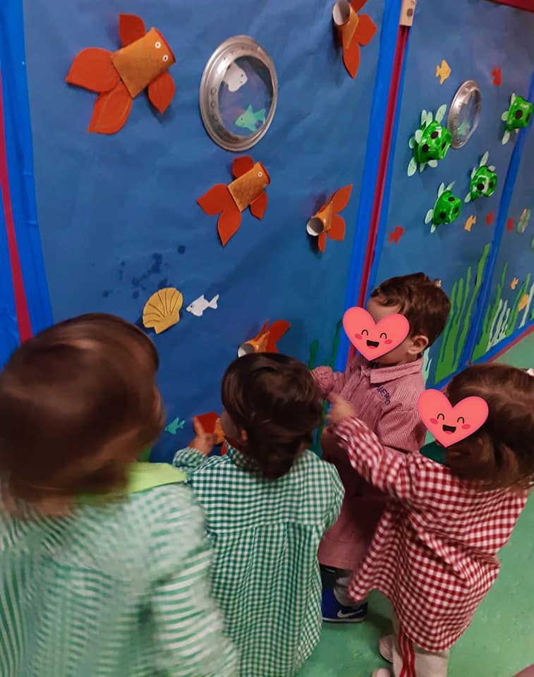 Un grupo de niños mira una pared con peces hechos con vasos de papel.
