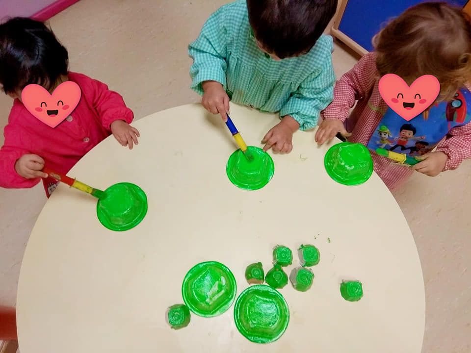 Tres niños están sentados en una mesa pintando círculos verdes con pinceles.