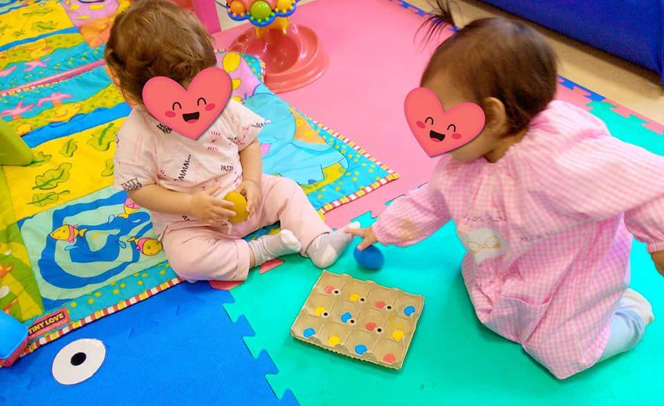 Dos bebés están sentados en una alfombra verde jugando con juguetes.