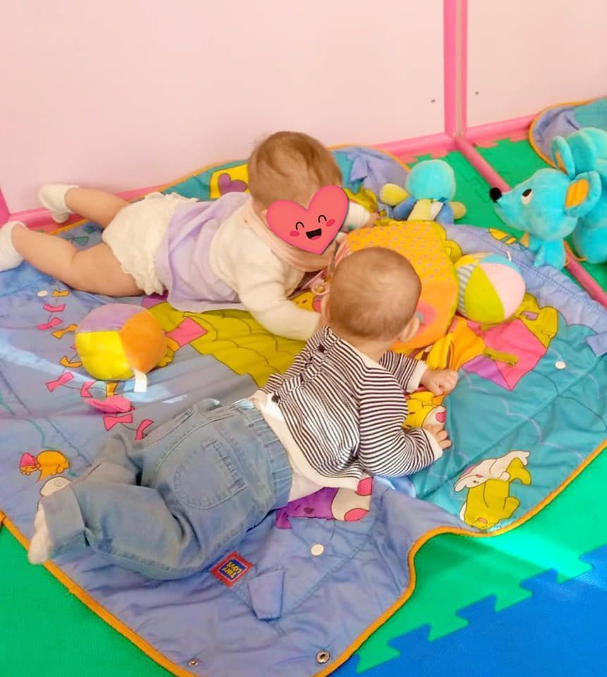 Dos bebés están acostados sobre una manta con juguetes y uno tiene un corazón en la cabeza.