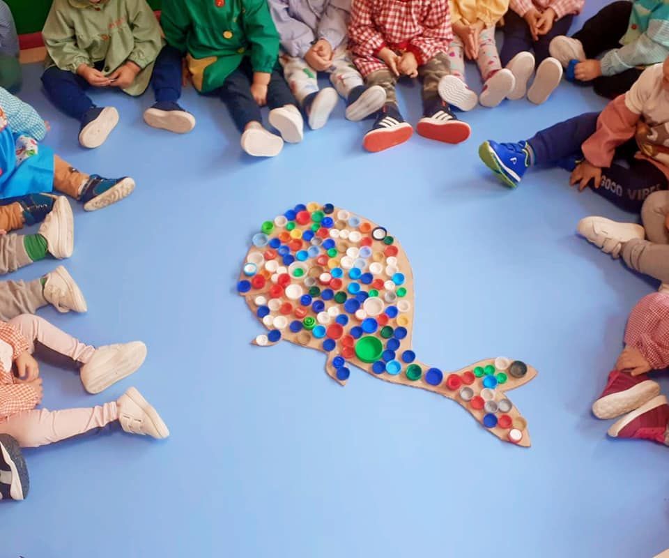 Un grupo de niños están sentados alrededor de una ballena hecha con tapas de botellas de plástico.