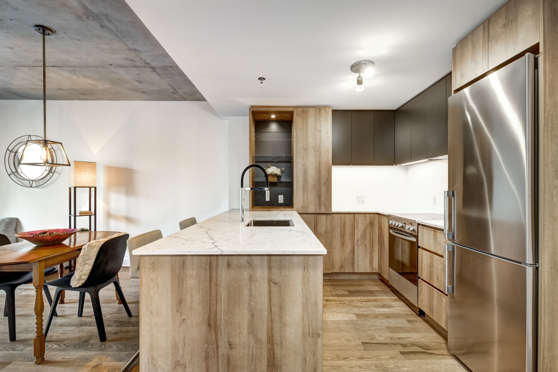 Cuisine moderne avec îlot en bois, réfrigérateur en inox et coin repas avec suspension lumineuse.