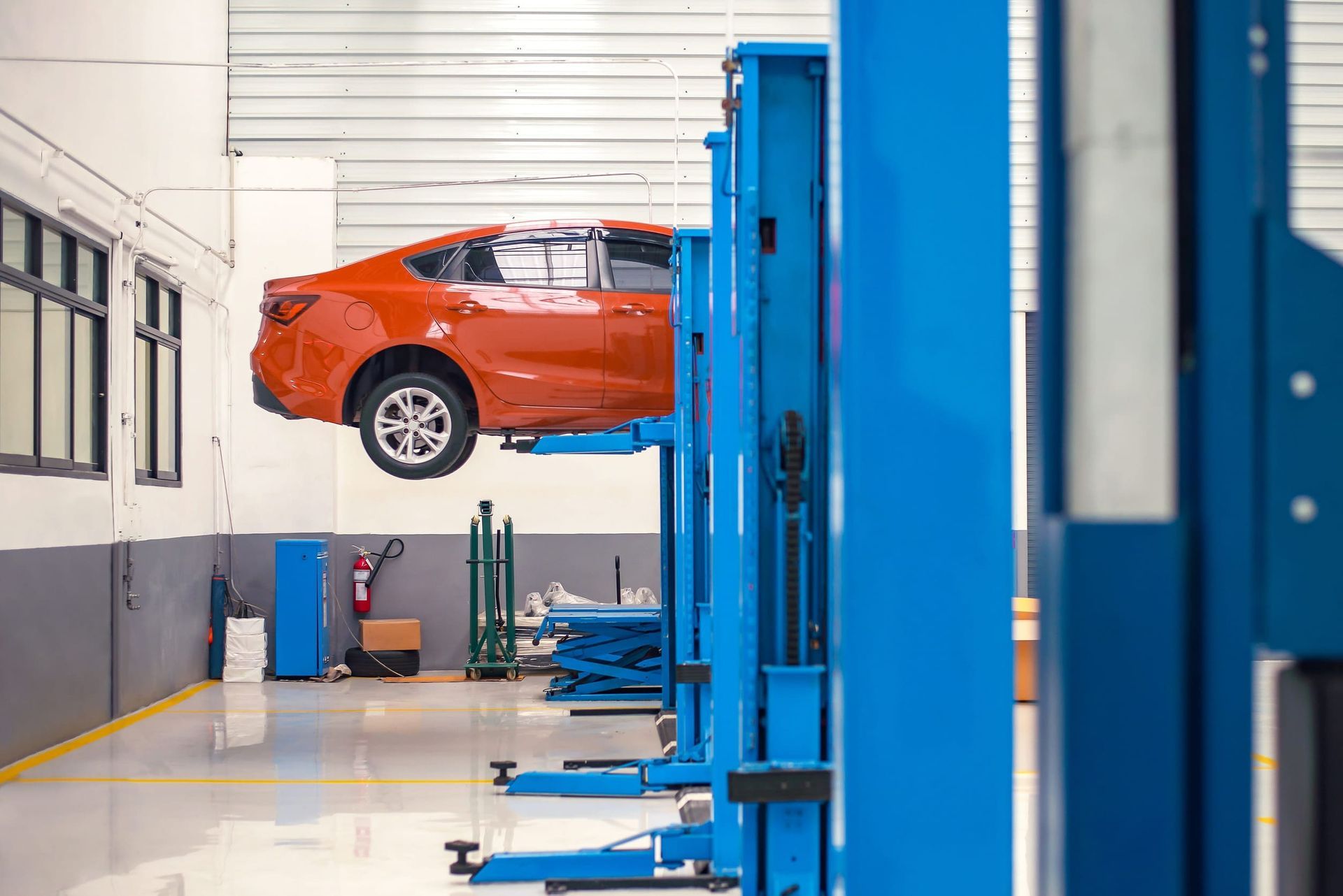 Un sedán naranja cuelga suspendido en el aire mediante un elevador automotriz azul dentro de un taller limpio y moderno.