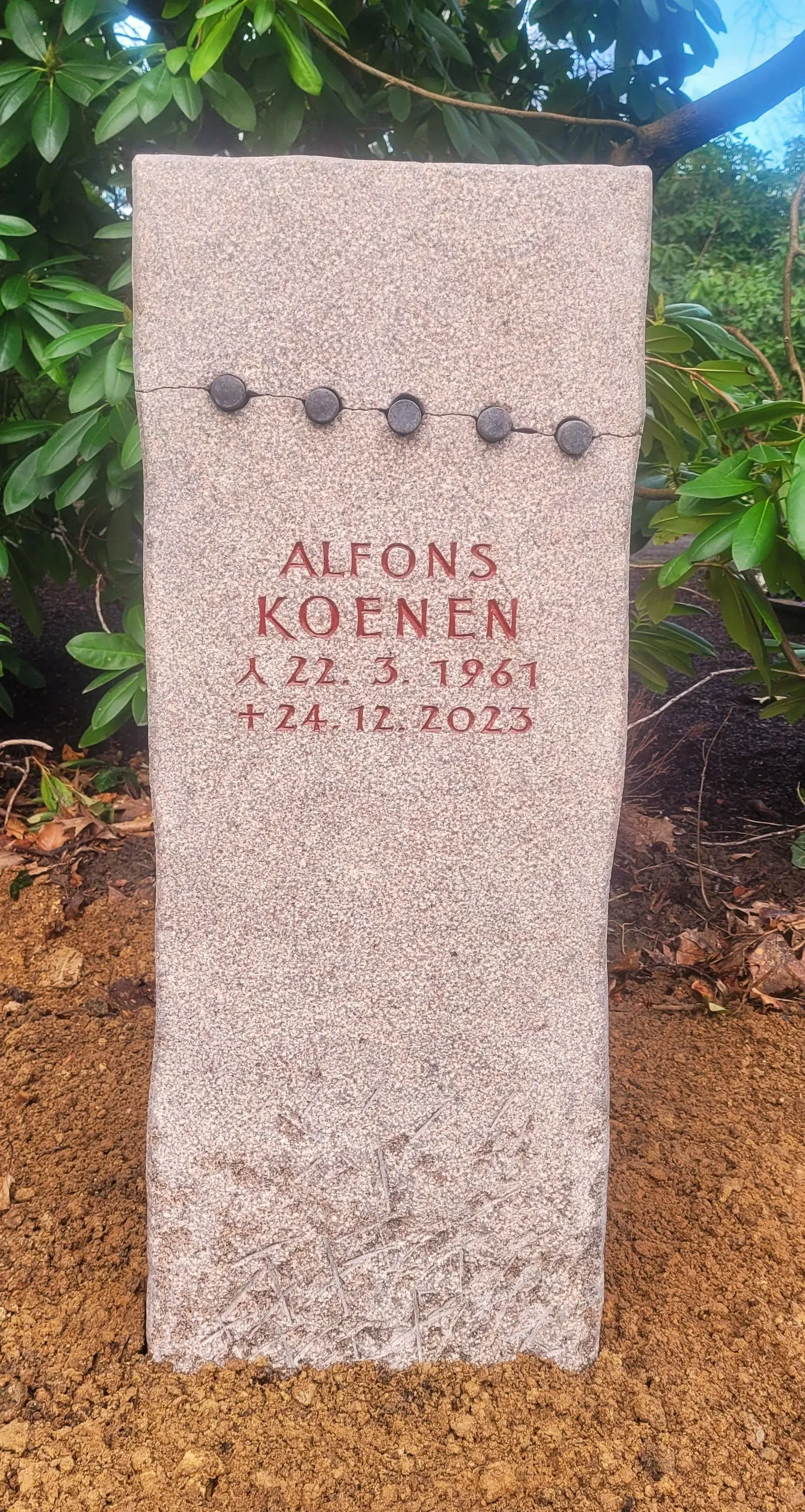 Helle Stele aus grobem Naturstein mit einer horizontalen Reihe runder Metallverzierungen.