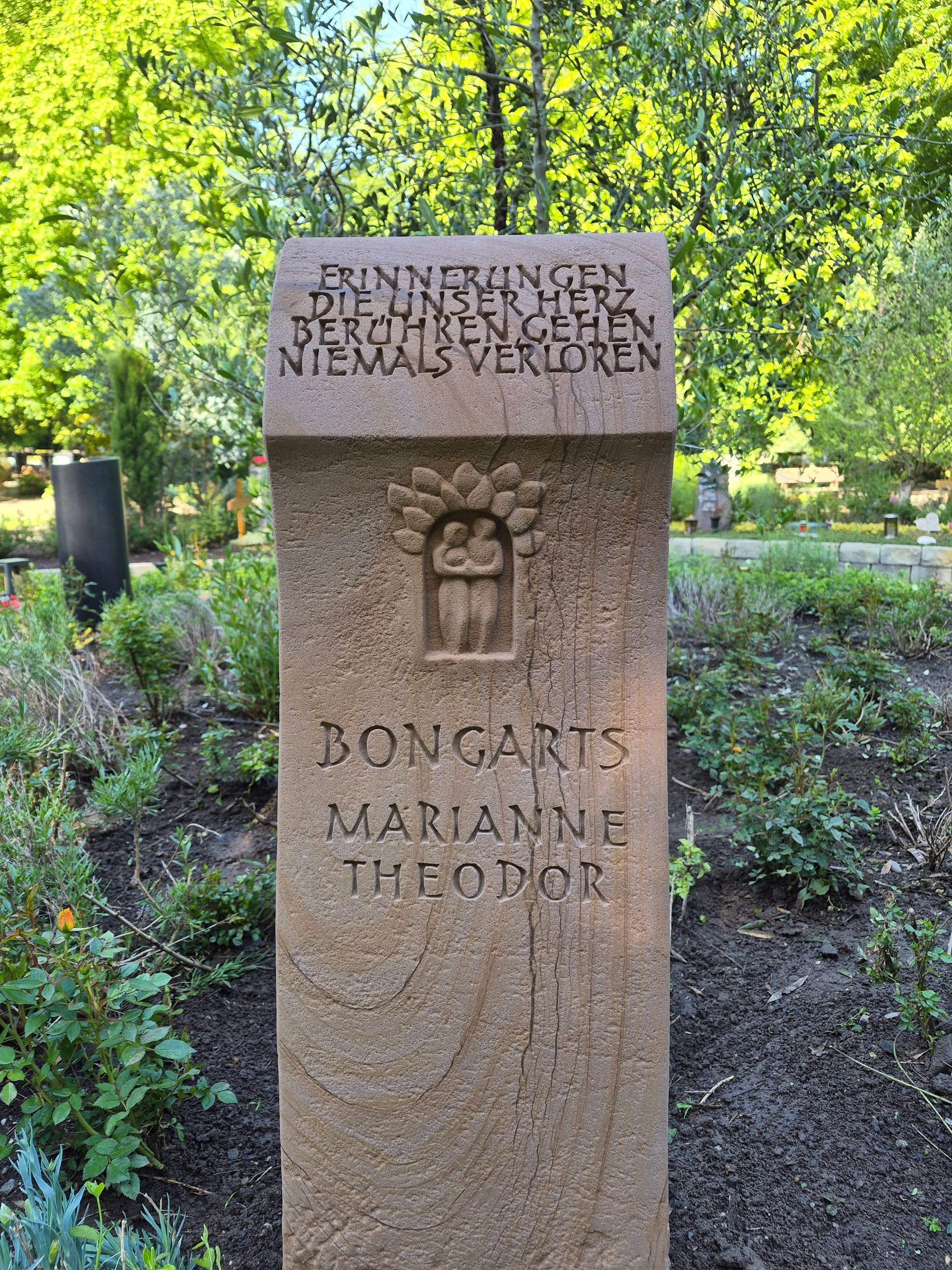 Verwittertes Steindenkmal mit deutscher Inschrift und kleiner geschnitzter Figur, umgeben von grünen Bäumen.