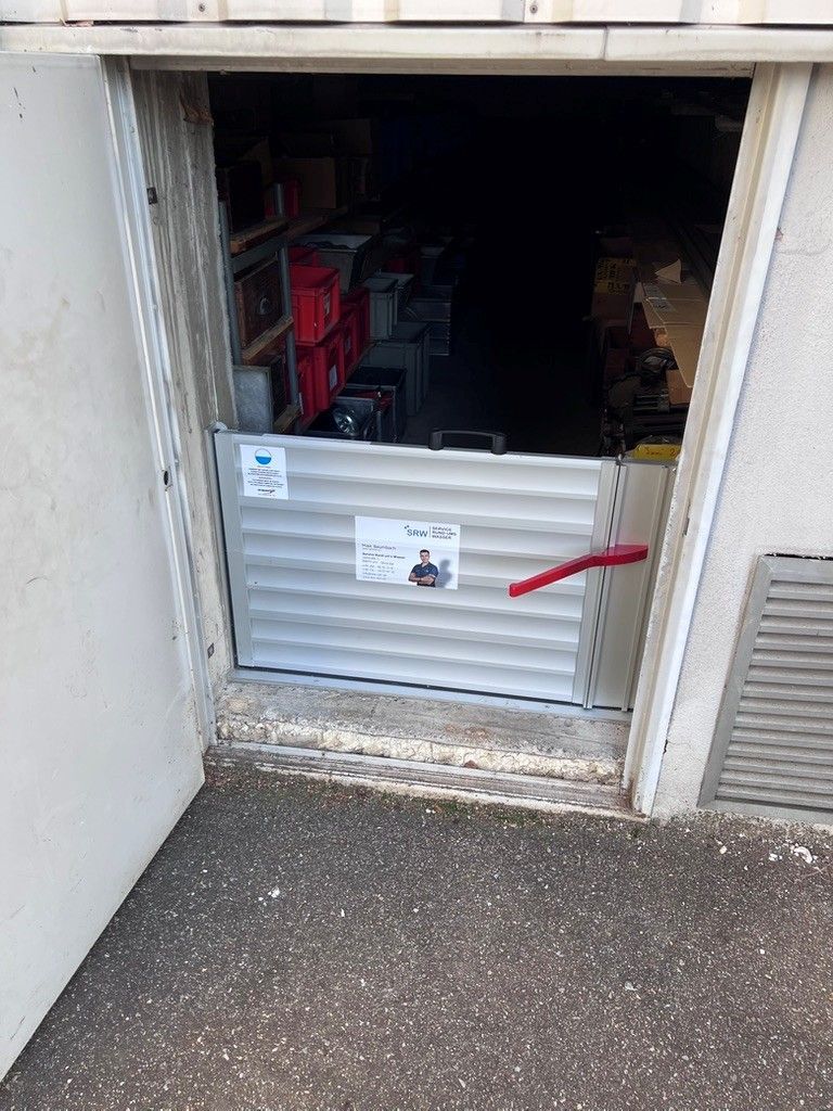 Hochwasserschutzbarriere in einem Türrahmen installiert. Silberne Platte mit rotem Griff, die einen dunklen Lagerraum schützt.