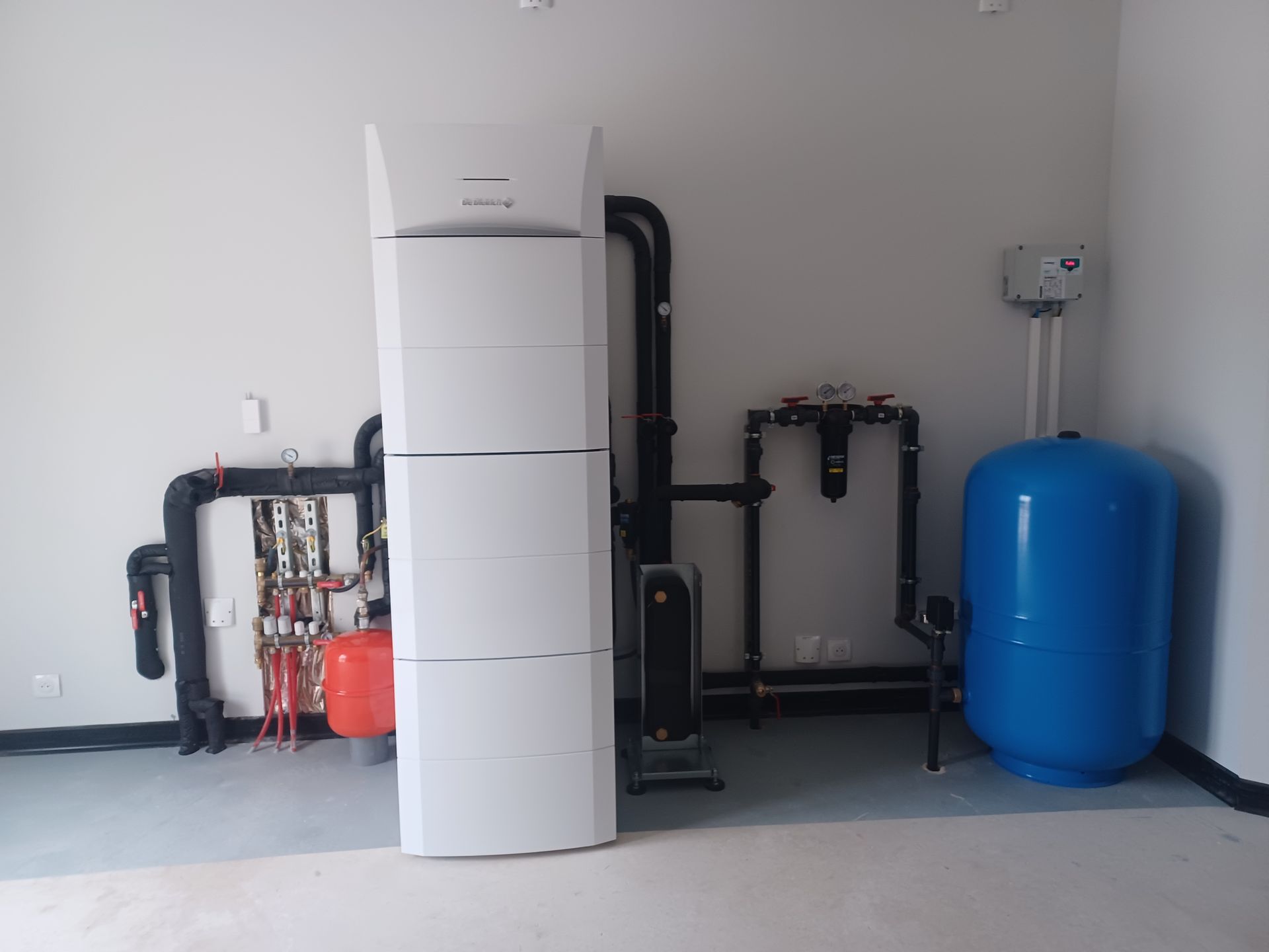 Pompe à chaleur blanche et plomberie associée dans une buanderie ; un réservoir de pression bleu et un vase d'expansion rouge sont également visibles.