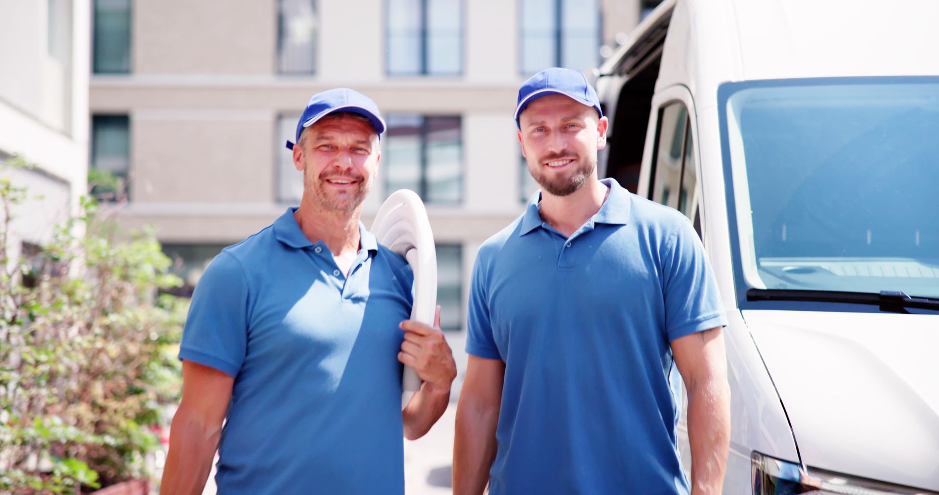 Deux hommes en chemises bleues et casquettes près d'une camionnette blanche, souriant à l'extérieur.