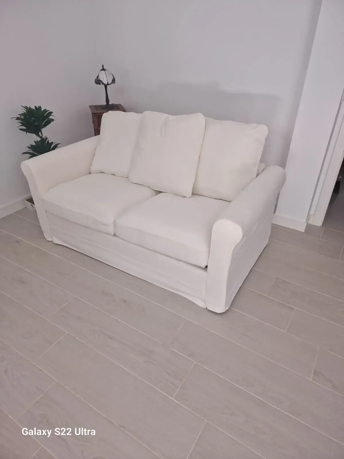 Sofá blanco de dos plazas con tres cojines en el respaldo en una habitación con suelo y paredes de color claro.