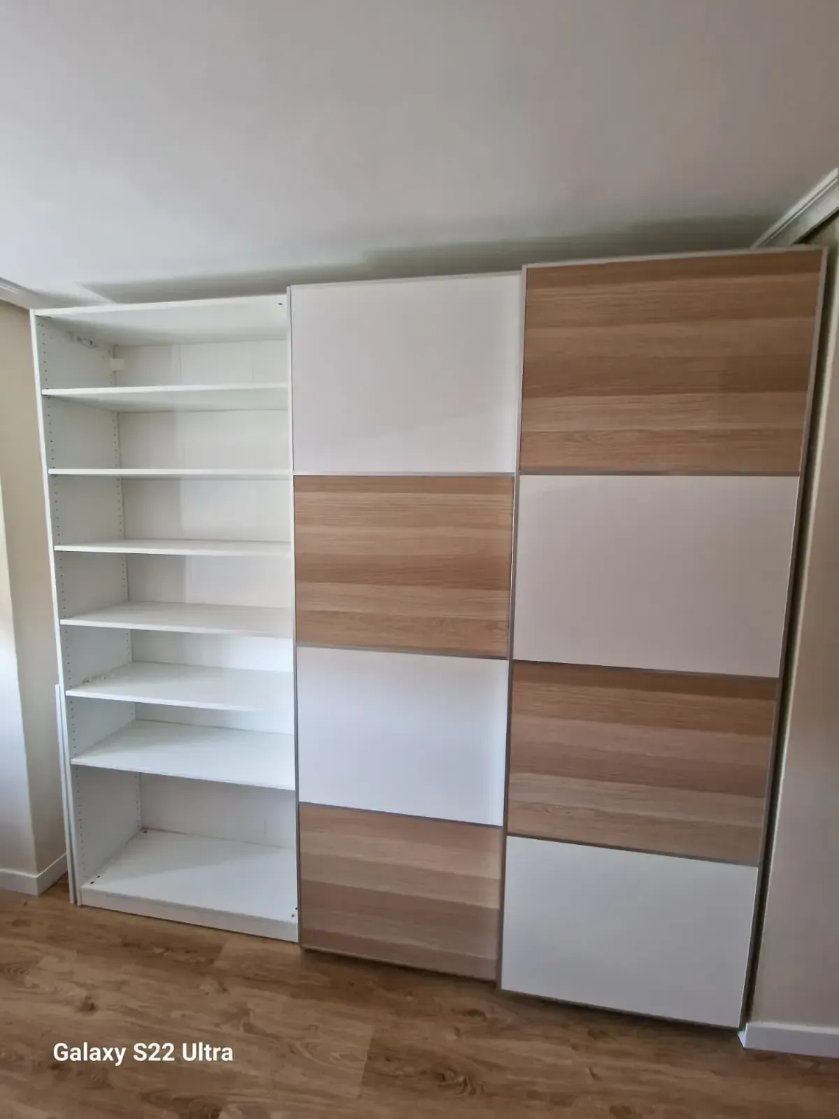 Armario con puertas correderas en tonos blancos y madera contra una pared blanca y con estanterías incorporadas.
