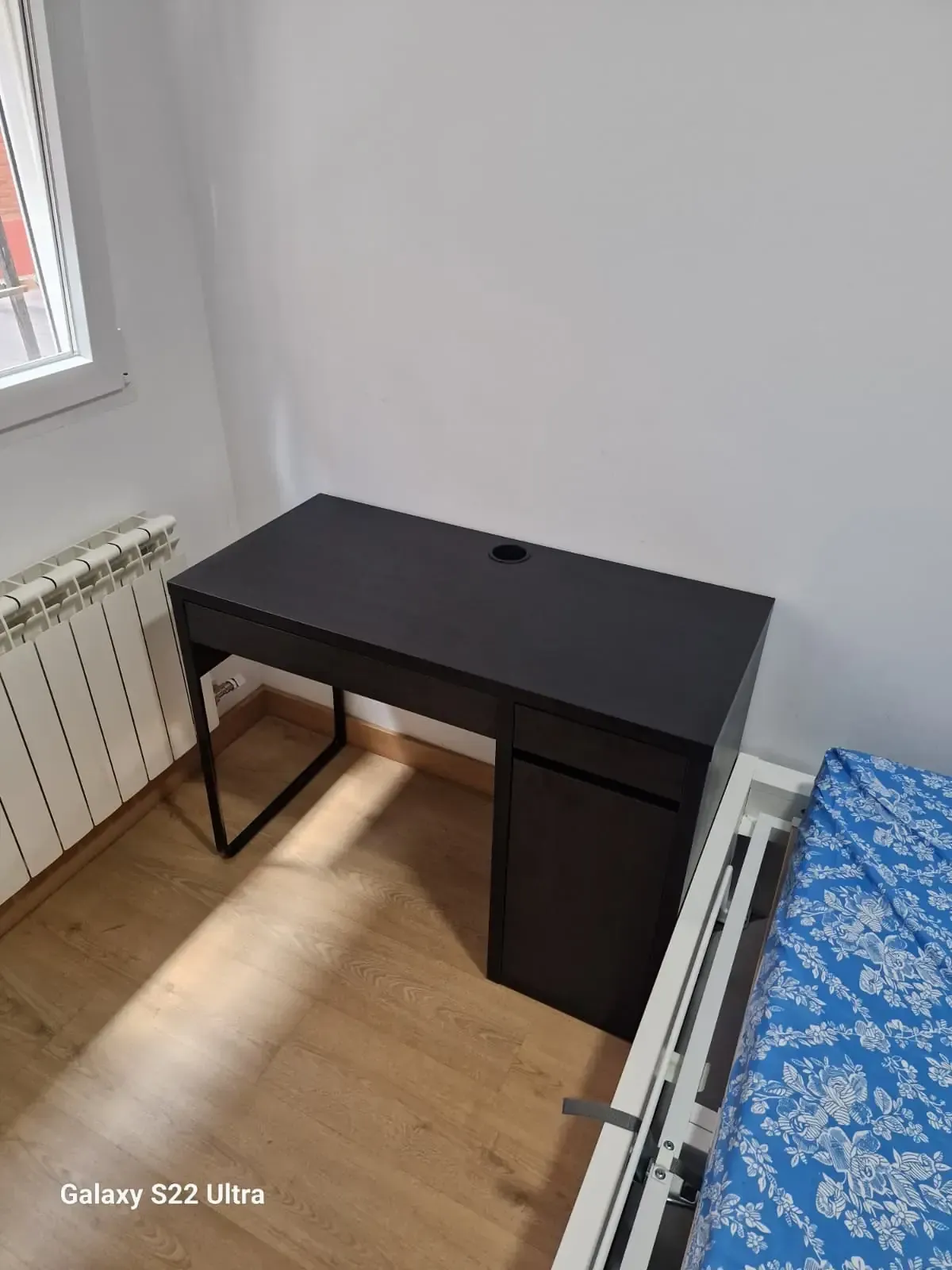 Escritorio negro contra una pared blanca en una habitación. A la izquierda hay un radiador y a la derecha una cama.